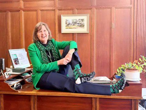 Person sitting on a desk in a green jacket and scarf, wearing socks with "1926" printed, with wood paneling in the background.