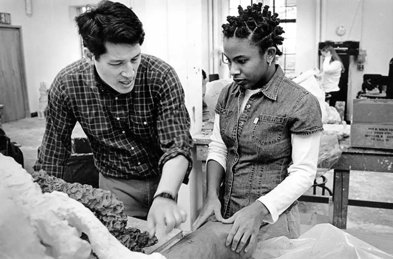 Two people working on a sculpture in a studio, one explaining while pointing, the other listening attentively.