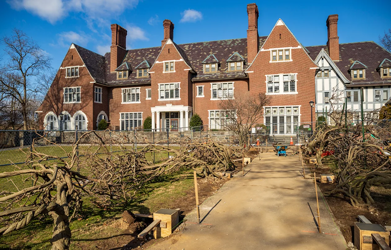 Large brick building with multiple chimneys and gabled roofs, set behind a path lined with trimmed trees and wooden benches on a sunny day.