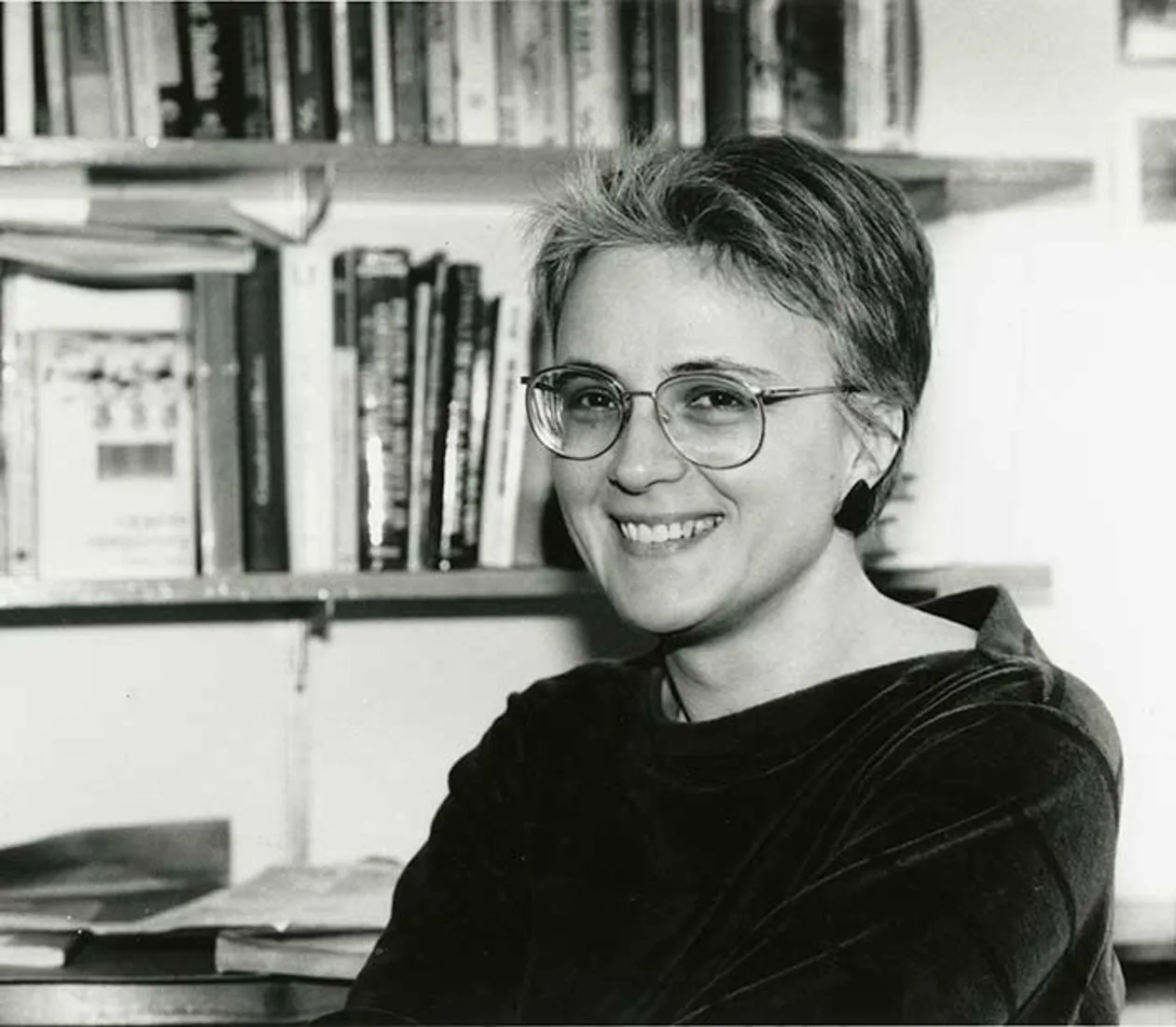 Person smiling, wearing glasses and sitting in front of a bookshelf filled with books.