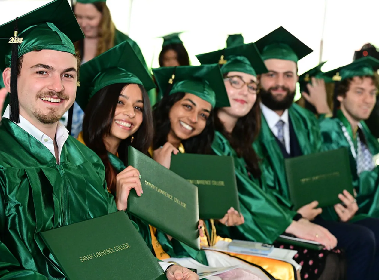 Graduates in green caps and gowns smile while holding diplomas at a ceremony for Sarah Lawrence College.