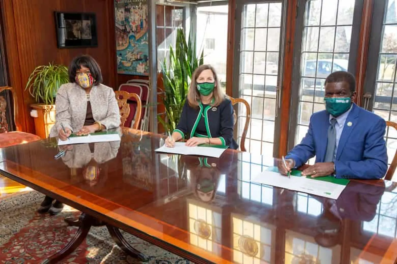 Three people wearing masks sit at a polished wooden table, signing documents in a bright room with large windows and plants.