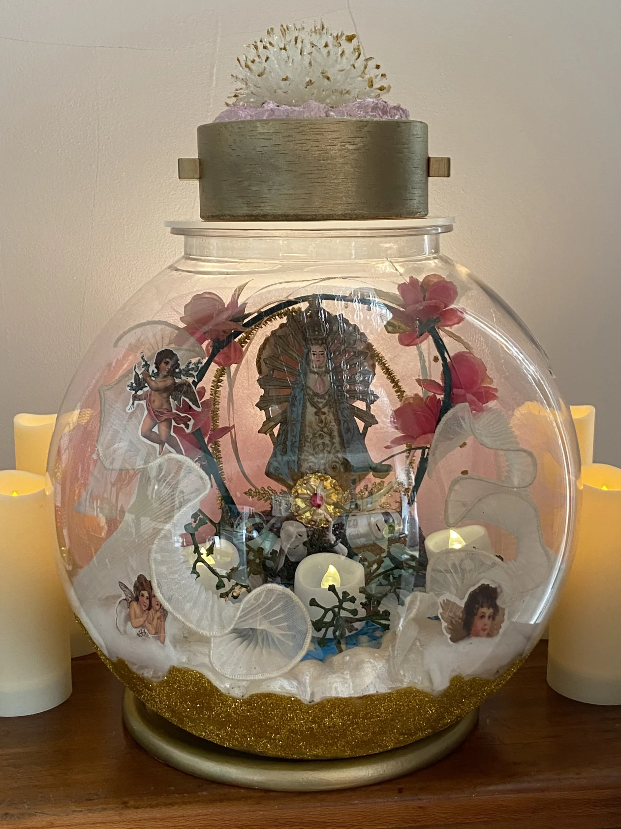 Decorative glass dome with religious figure, flowers, and angel figurines. Surrounded by candles, with a textured object on top.