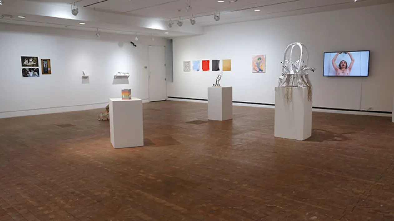 Art gallery with various artworks on walls, sculptures on pedestals, and a video screen displaying a person with raised arms.