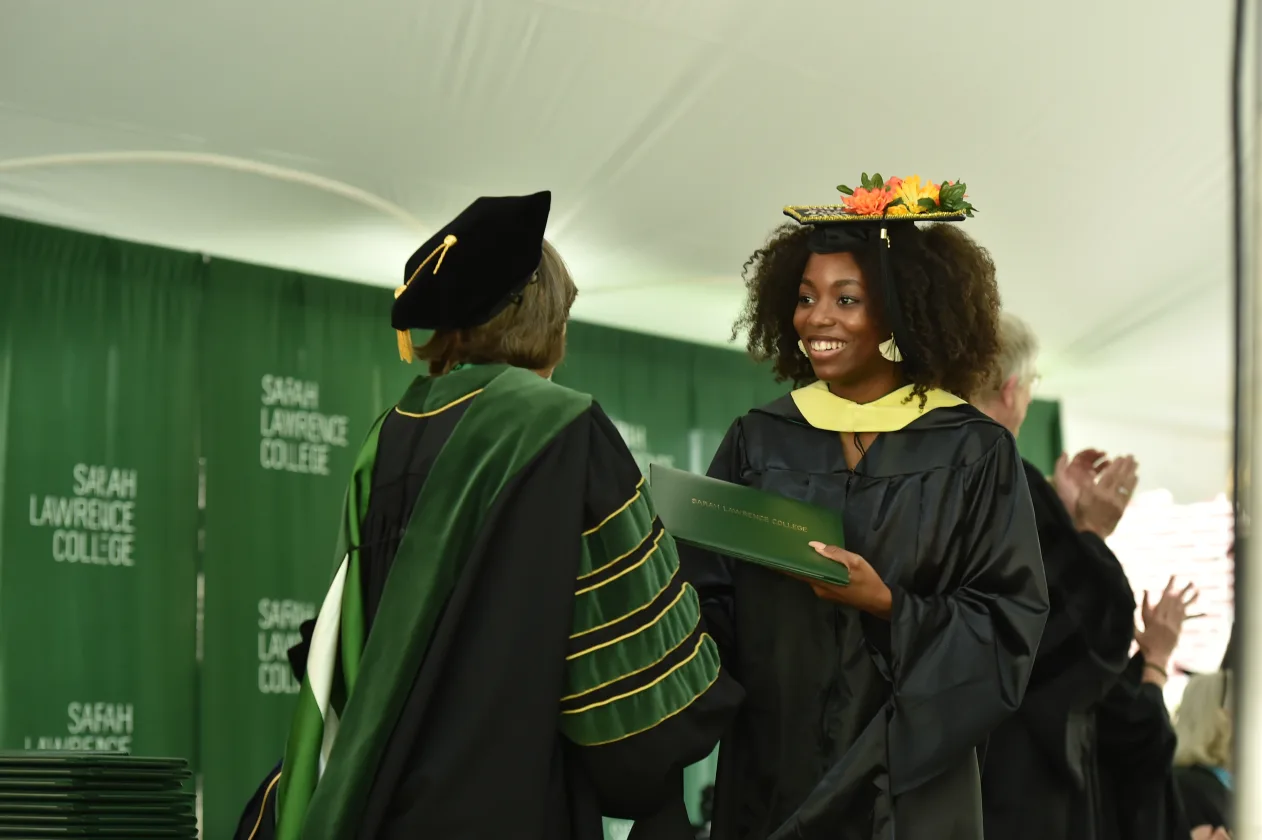 Graduate receiving diploma on stage, wearing a decorated cap and gown, with green Sarah Lawrence College banners in the background.