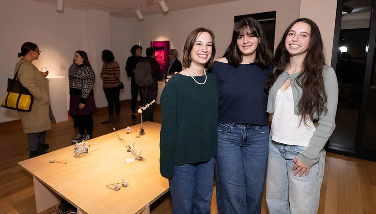 Three people standing together in a gallery, with art pieces on a table in front and others looking at exhibits in the background.