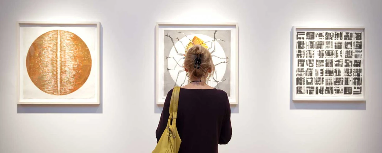 Person with a yellow bag views three abstract artworks in a gallery: a copper circle, a cracked sunburst, and a black-and-white pattern.