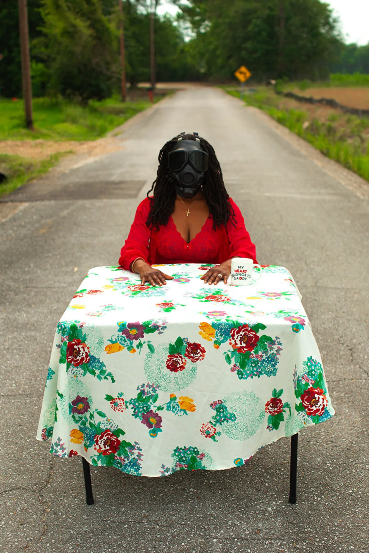 Person in a red shirt wearing a gas mask sits at a table with a flowery cloth, on a deserted road lined with trees.