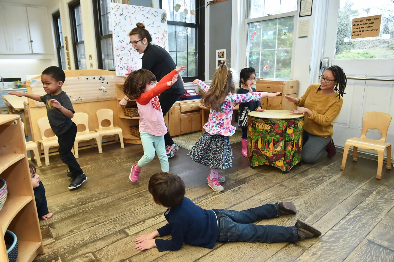 Children play and dance in a classroom with musical instruments and colorful decor. Two adults supervise the activity.
