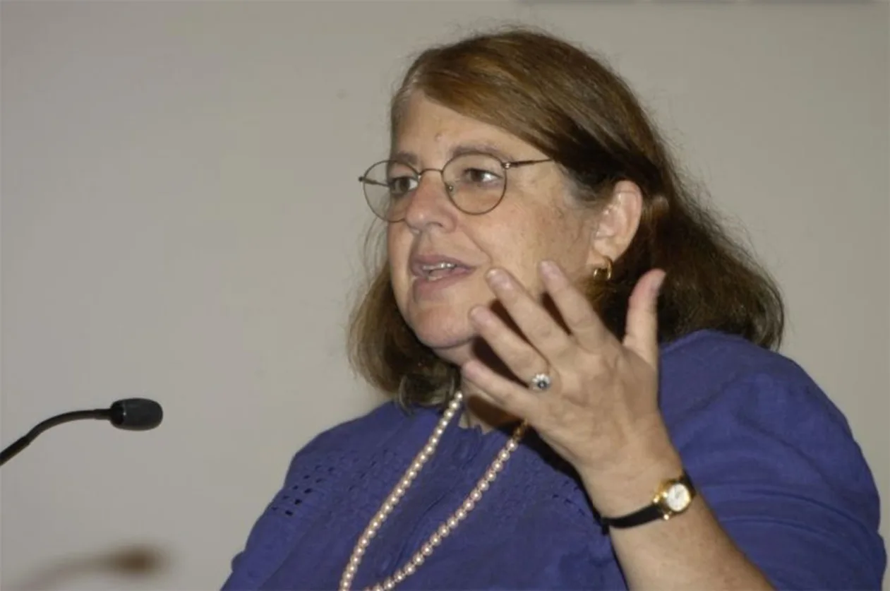 A person with glasses speaks into a microphone, gesturing with one hand. They are wearing a blue shirt and necklace.