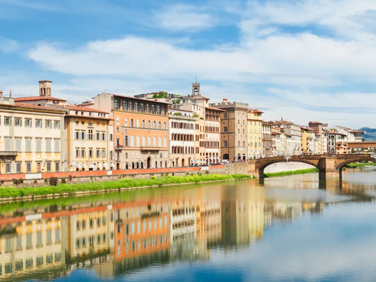 A row of colorful historic buildings with arched windows line a riverbank under a blue sky, reflecting in the calm water.