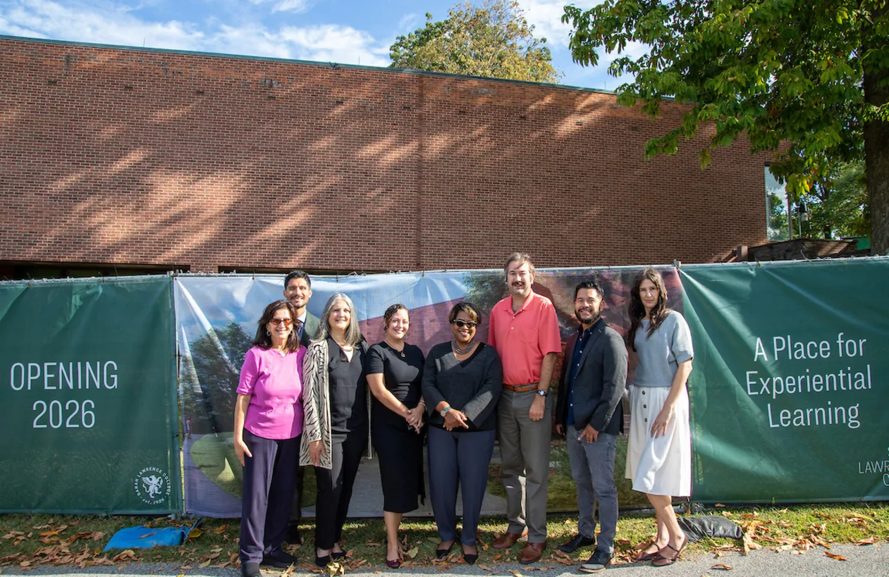 Group of nine people standing in front of a brick building with banners displaying "Opening 2026" and "A Place for Experiential Learning."