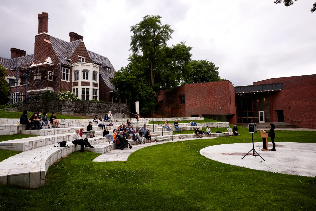 Outdoor event at an amphitheater with an audience seated on tiered steps facing a speaker at a podium. Brick and stone buildings in the background.