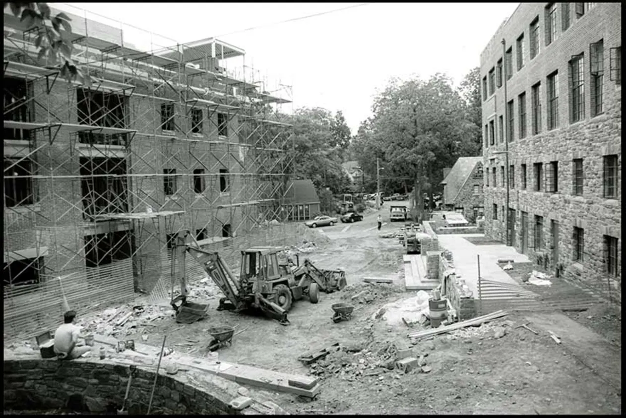 Construction site with scaffolding on a building's facade, a backhoe, and scattered materials. Trees and more buildings in the background.