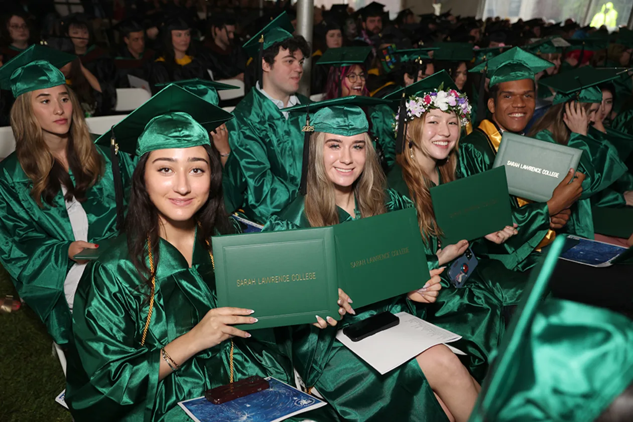 Graduates in green caps and gowns sit in rows, holding diplomas and smiling, during a ceremony.