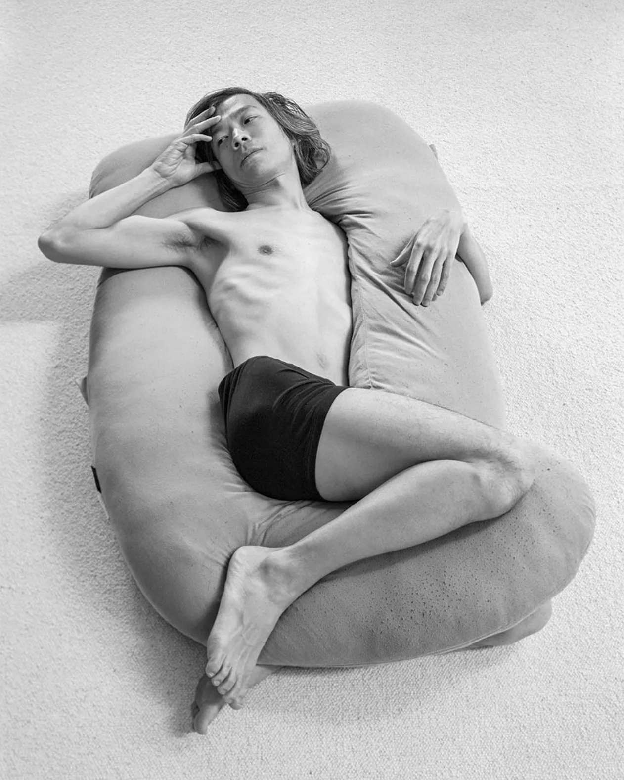 Person reclining on a large pillow, hand on forehead, wearing dark shorts, in a relaxed pose on a light-colored carpet.