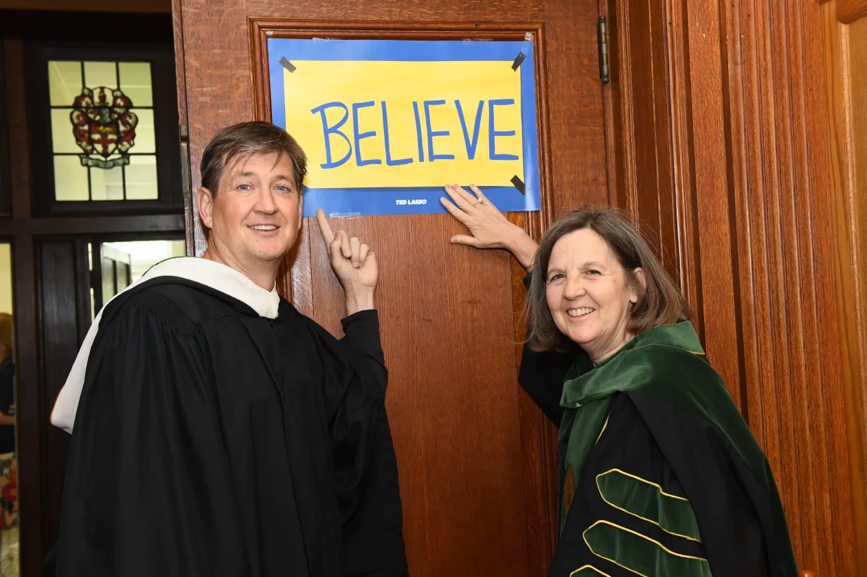 Two people in academic robes smiling and pointing at a sign on a wooden door that reads "BELIEVE" in blue letters on a yellow background.