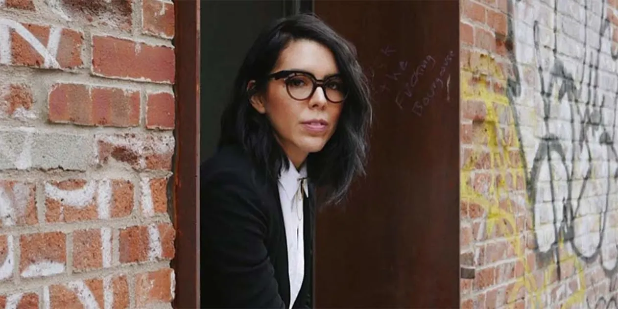 Person with glasses and dark hair leans out of a doorway between two graffiti-covered brick walls, wearing a black jacket and white shirt.