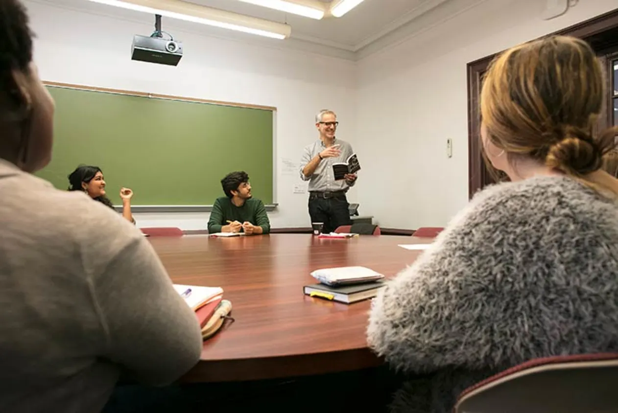 Person stands at the head of a table in a classroom, holding a book, while several people sit around listening attentively.