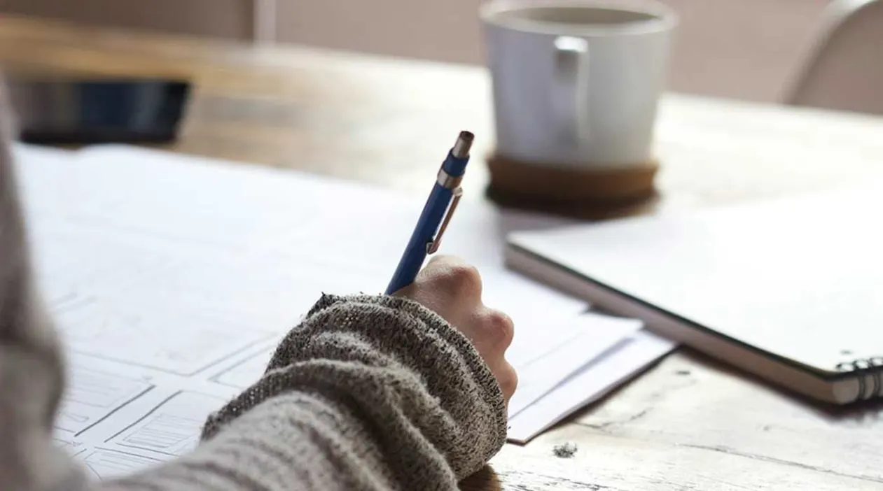 Person writing on paper at a wooden table with a pen, wearing a sweater. A cup and notebook are on the table in the background.