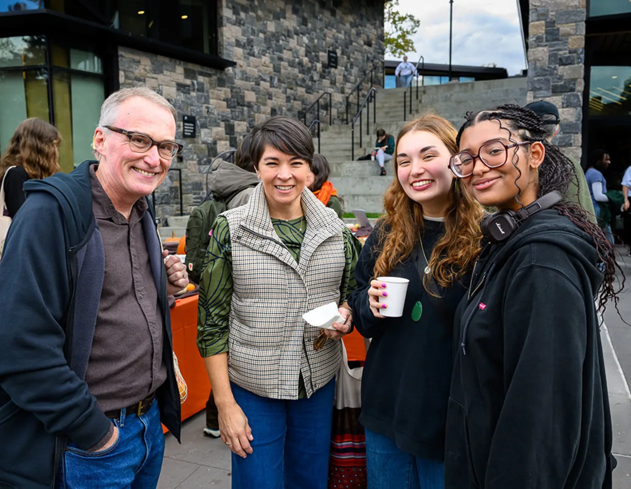 Four people smiling at an outdoor event, standing in front of a stone building. One holds a cup, and another has headphones around their neck.