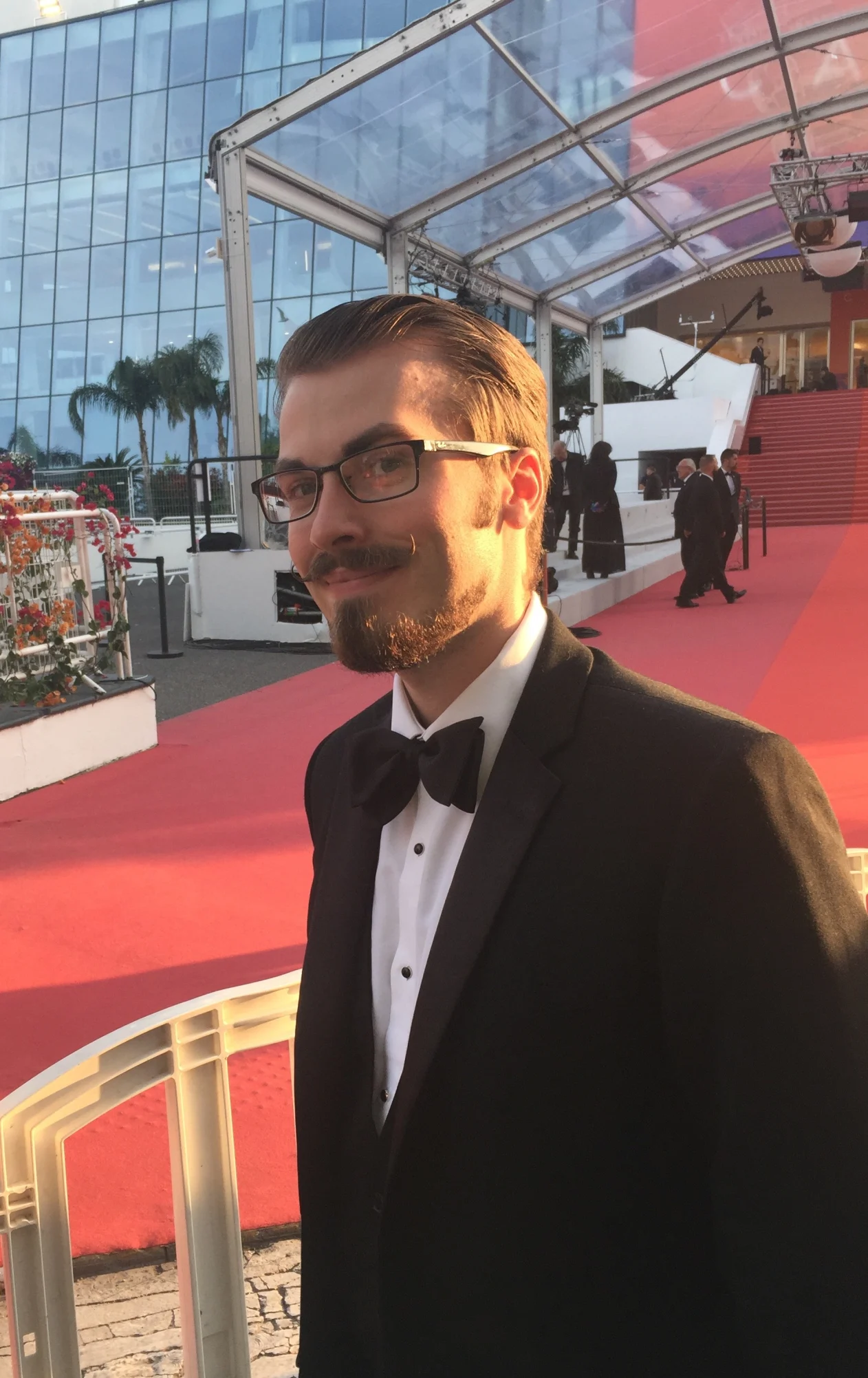 Person in a tuxedo stands on a red carpet under a clear canopy, with people and stairs in the background.