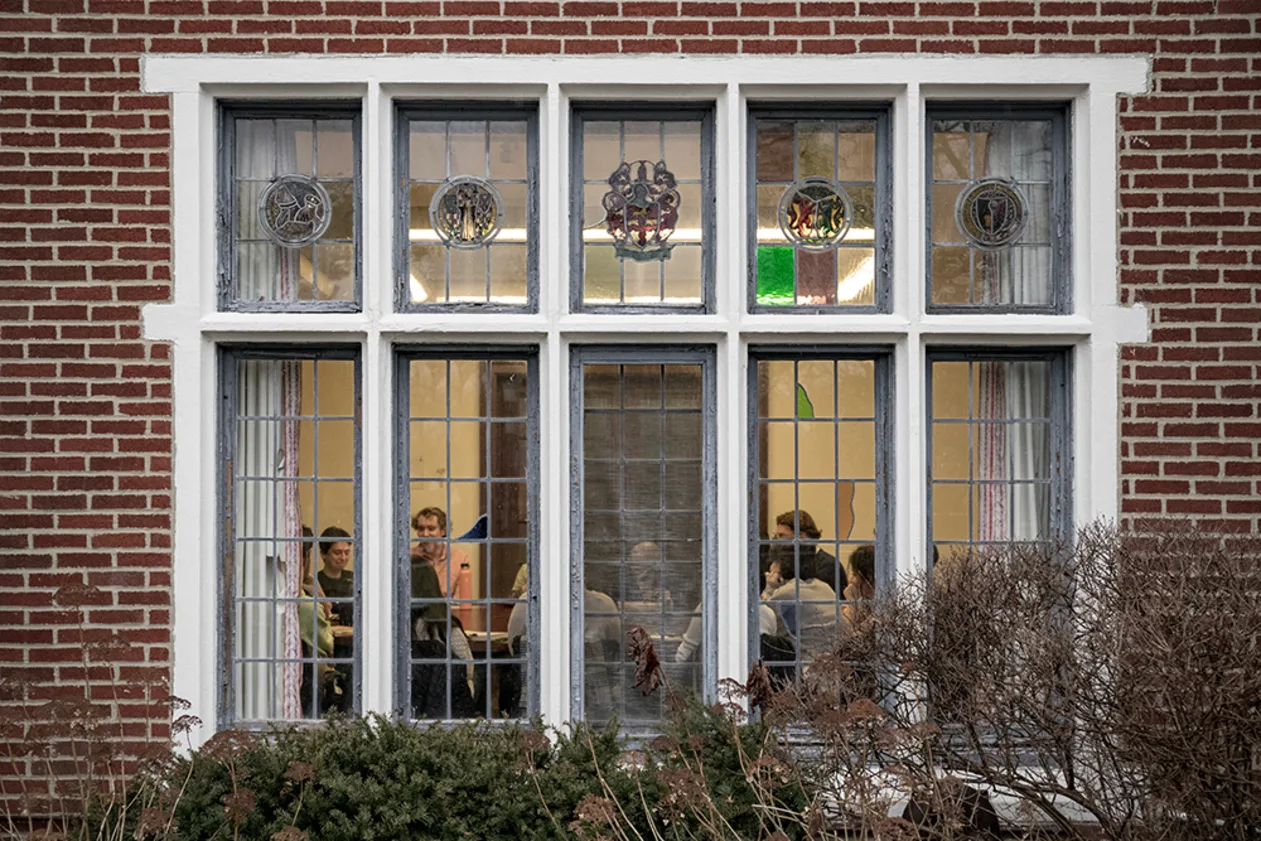 People seen through large, multi-paneled window of brick building, engaged in discussion. Stained glass details visible above the window.