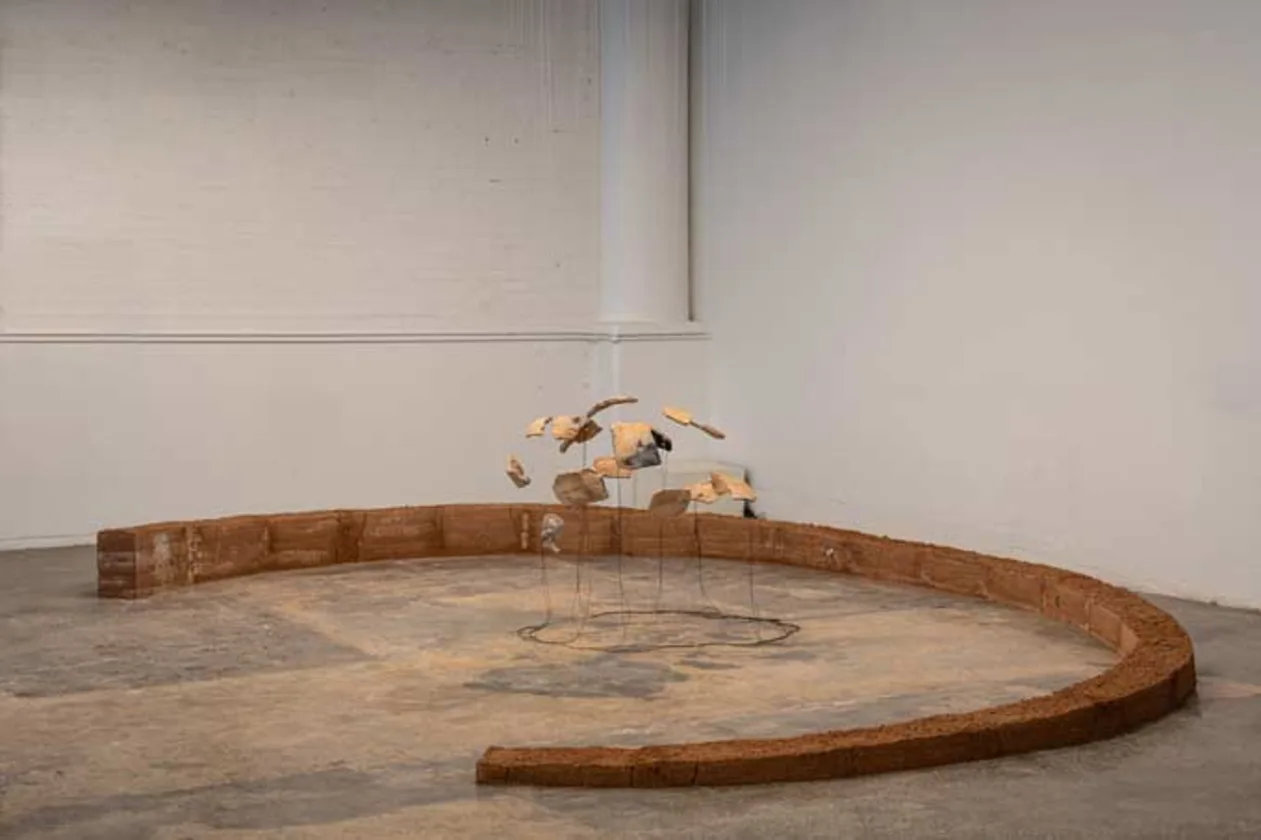 Art installation with suspended rocks and clay bricks forming a semi-circular wall on a concrete floor against a white gallery wall.