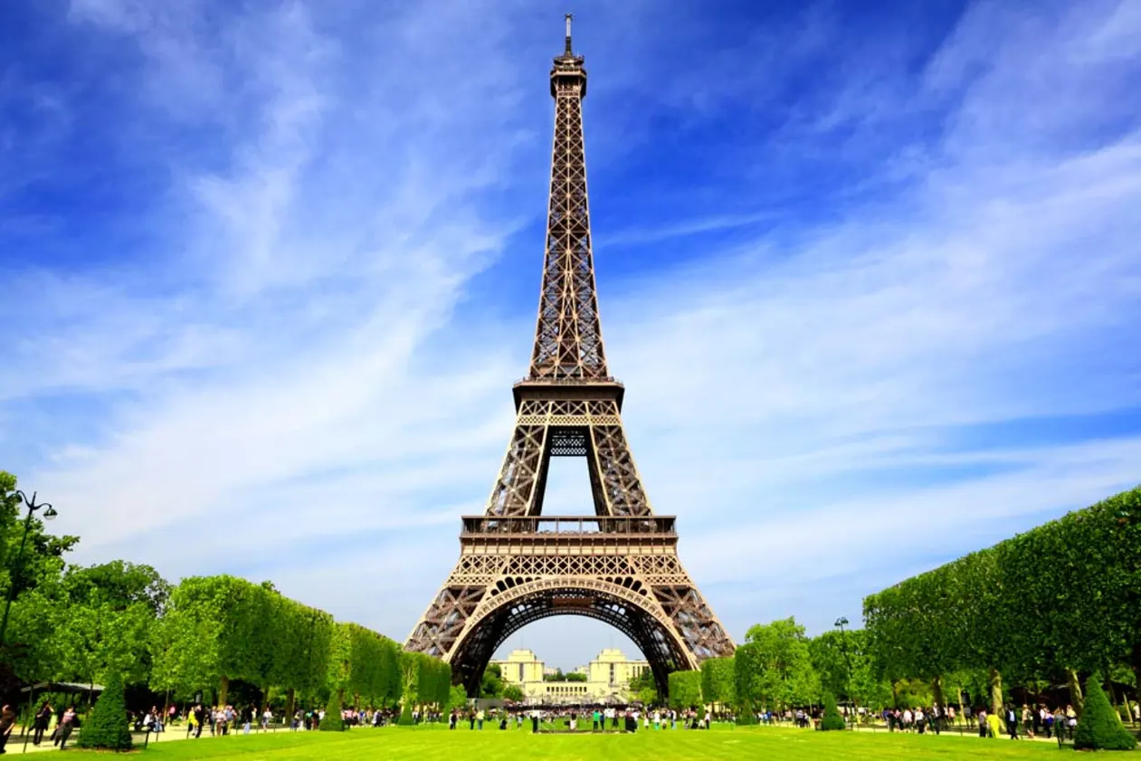 The Eiffel Tower in Paris under a bright blue sky with scattered clouds, surrounded by green trees and a grass field with people.