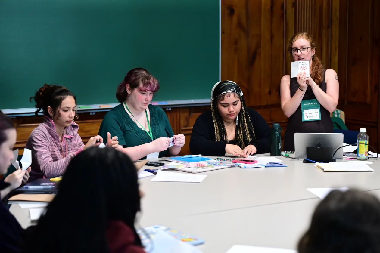 A group of people sits around a table with papers and laptops, while one person stands and speaks, holding a document.