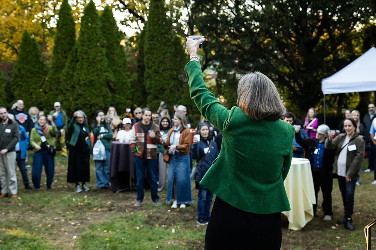 Person in green jacket raises a glass to a group of people outdoors, with trees and a tent in the background.