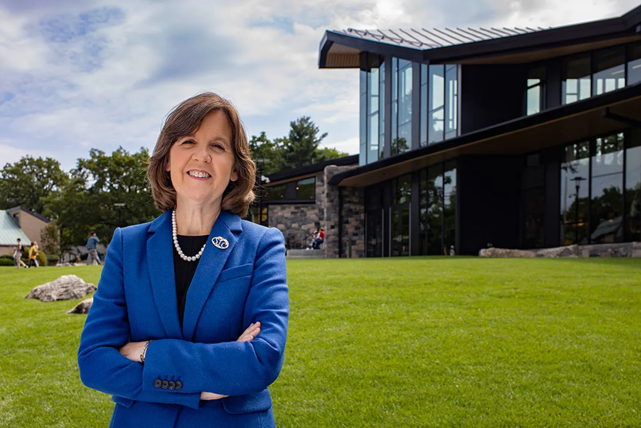 Person in a blue suit stands confidently on a grassy lawn in front of a modern building with large windows and a sloped roof.