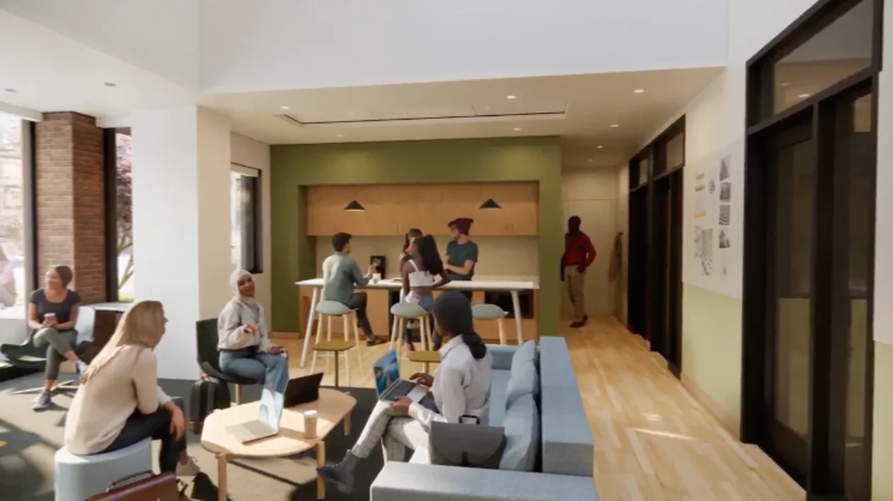 People in a modern lounge, sitting on sofas and stools, engaging with laptops and talking. Bright space with large windows and light wood flooring.
