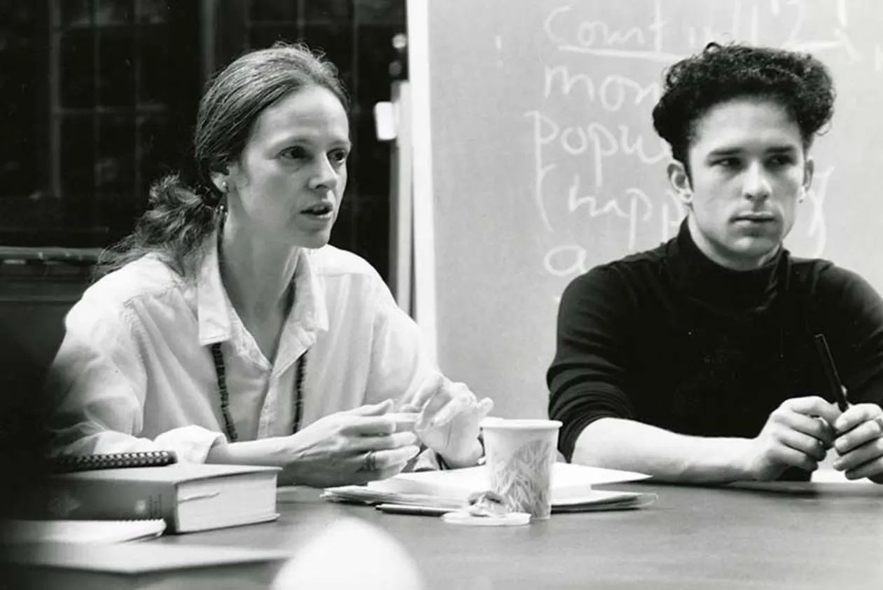 Two people sit at a table with books and papers, discussing something. A chalkboard with partial writing is in the background.