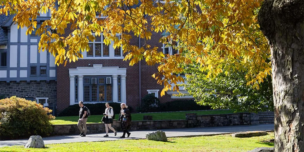 Three people walk along a path in front of a brick building, under a tree with autumn leaves.