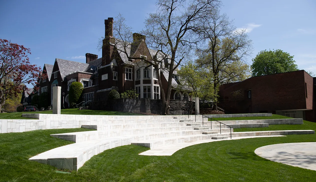 Historic brick building with tall chimneys surrounded by trees and manicured lawns, featuring modern tiered seating in the foreground.