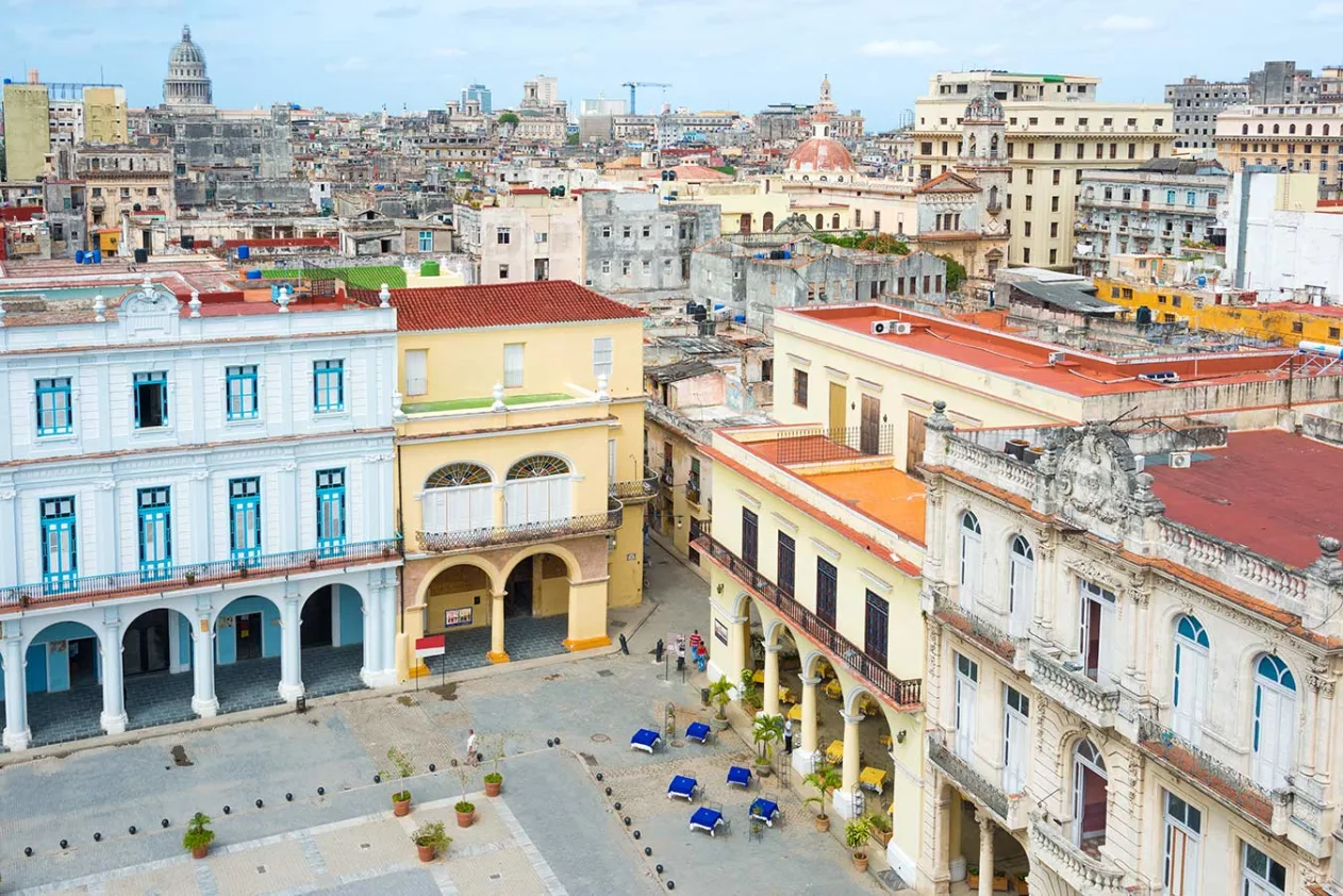 A vibrant cityscape with colorful colonial buildings surrounding a plaza, featuring classic architecture and distant skyline views.