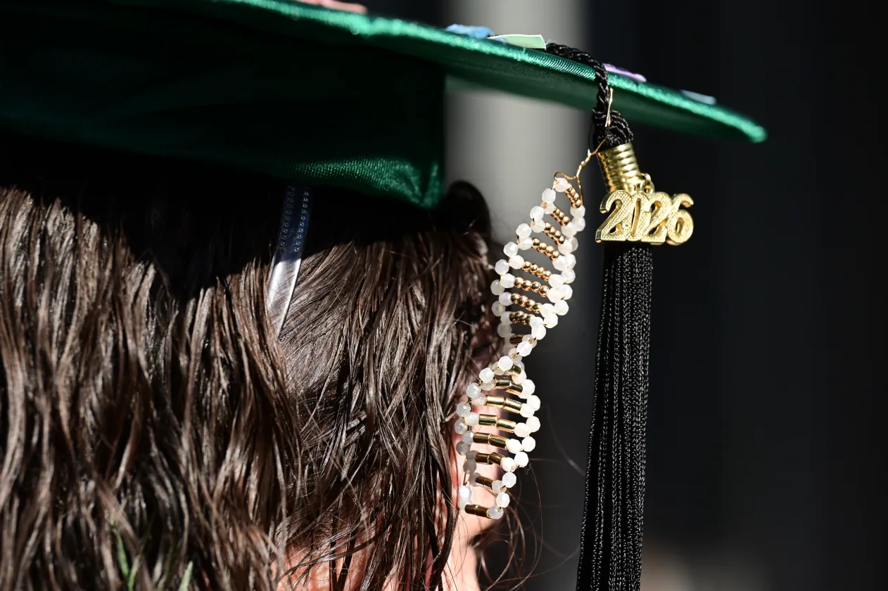 Green graduation cap with DNA-shaped decoration and a tassel featuring "2026" in gold, worn by a person with wet hair.