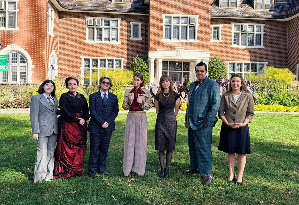 Seven people in vintage clothing stand on grass in front of a large brick building with white-trimmed windows and green bushes.