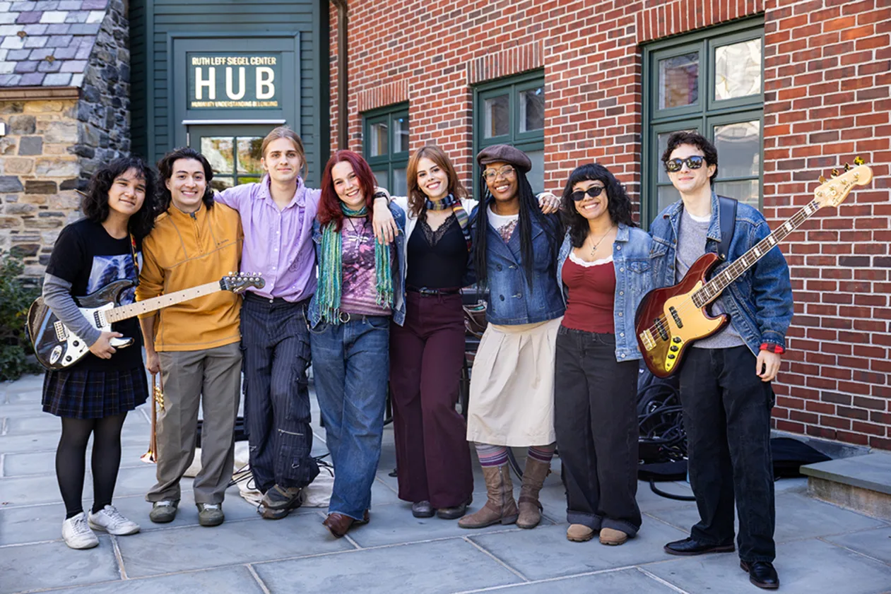 Eight people stand together smiling, some holding guitars, in front of a brick building with a "Hub" sign. Casual attire, outdoors.