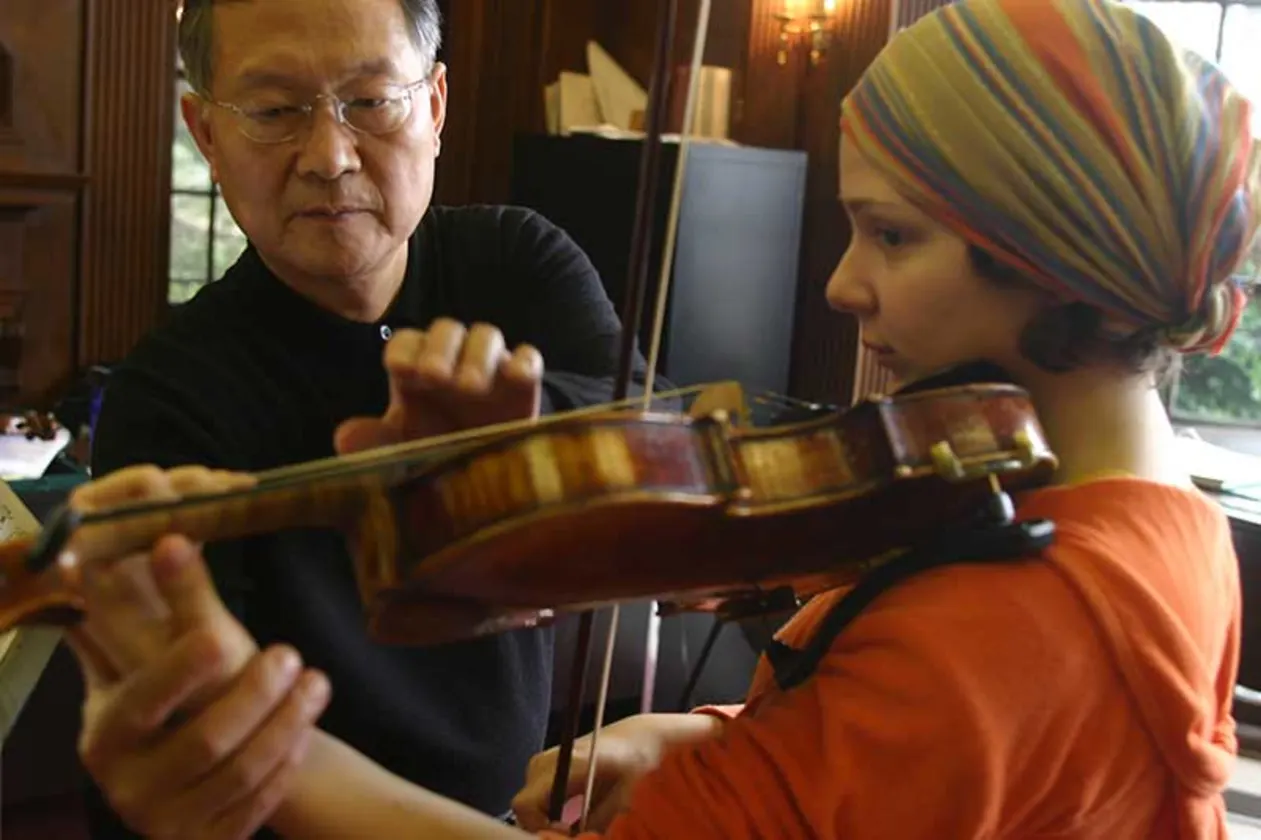 Person assists another in adjusting a violin posture, indoors with wood-paneled walls. The second person wears an orange top and colorful headscarf.