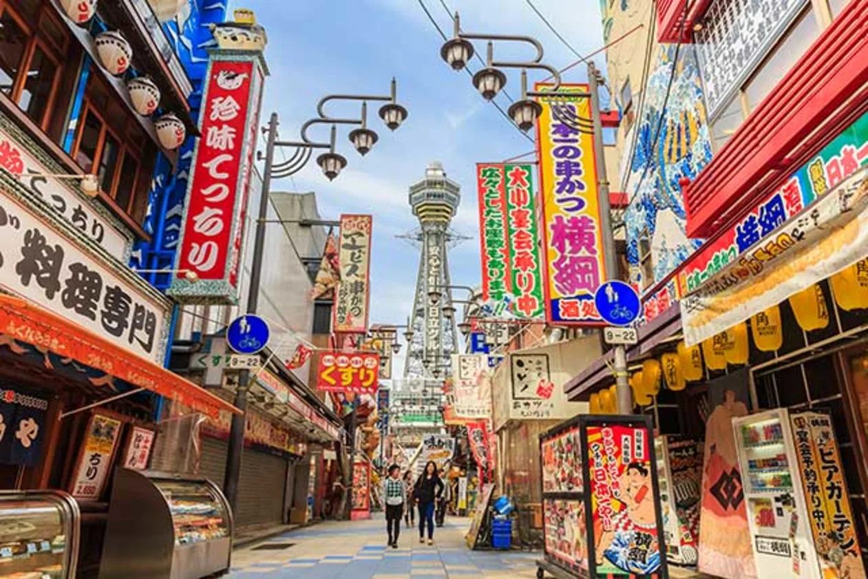 Bustling street in Japan with colorful signs, lanterns, and a prominent tower in the background. People walk along the lively pathway.
