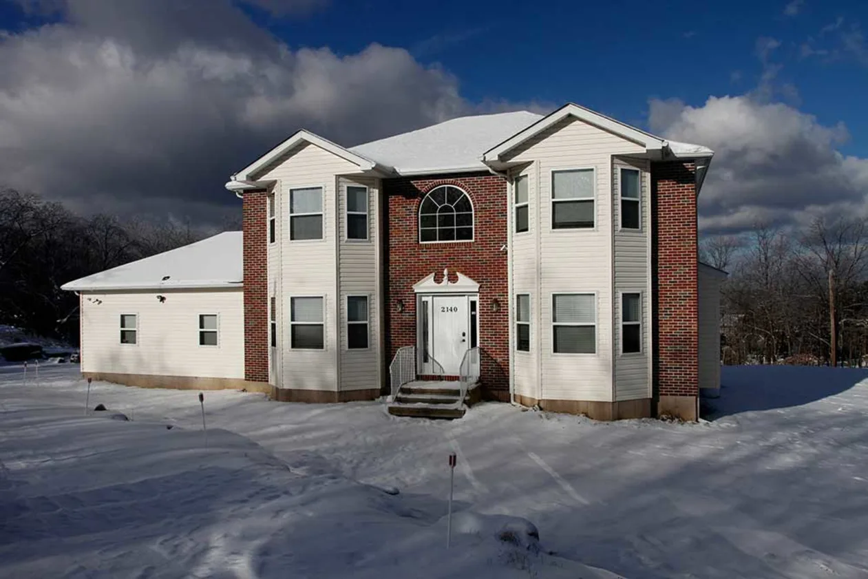 A two-story house with red brick and white siding stands in a snowy landscape under a partly cloudy sky.