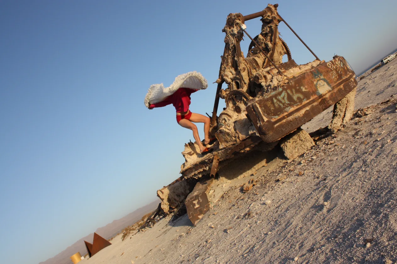 Person in a red outfit leaps from a decaying, rusted machine in a desert landscape under a clear blue sky.