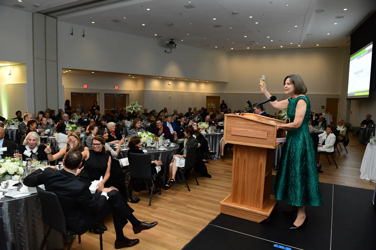 Speaker in green attire raises a glass at a podium in a banquet hall filled with seated attendees at round tables.