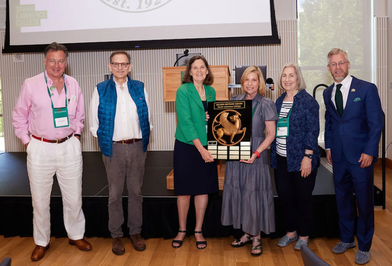 Six individuals stand on a stage, with two holding an award plaque with a decorative design. A podium and screen are visible in the background.