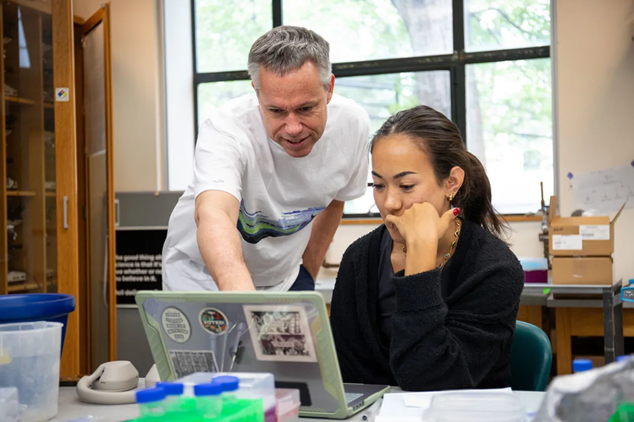 Two people in a room focus on a laptop. One points at the screen while the other sits, looking thoughtful. Shelves and lab items are in the background.