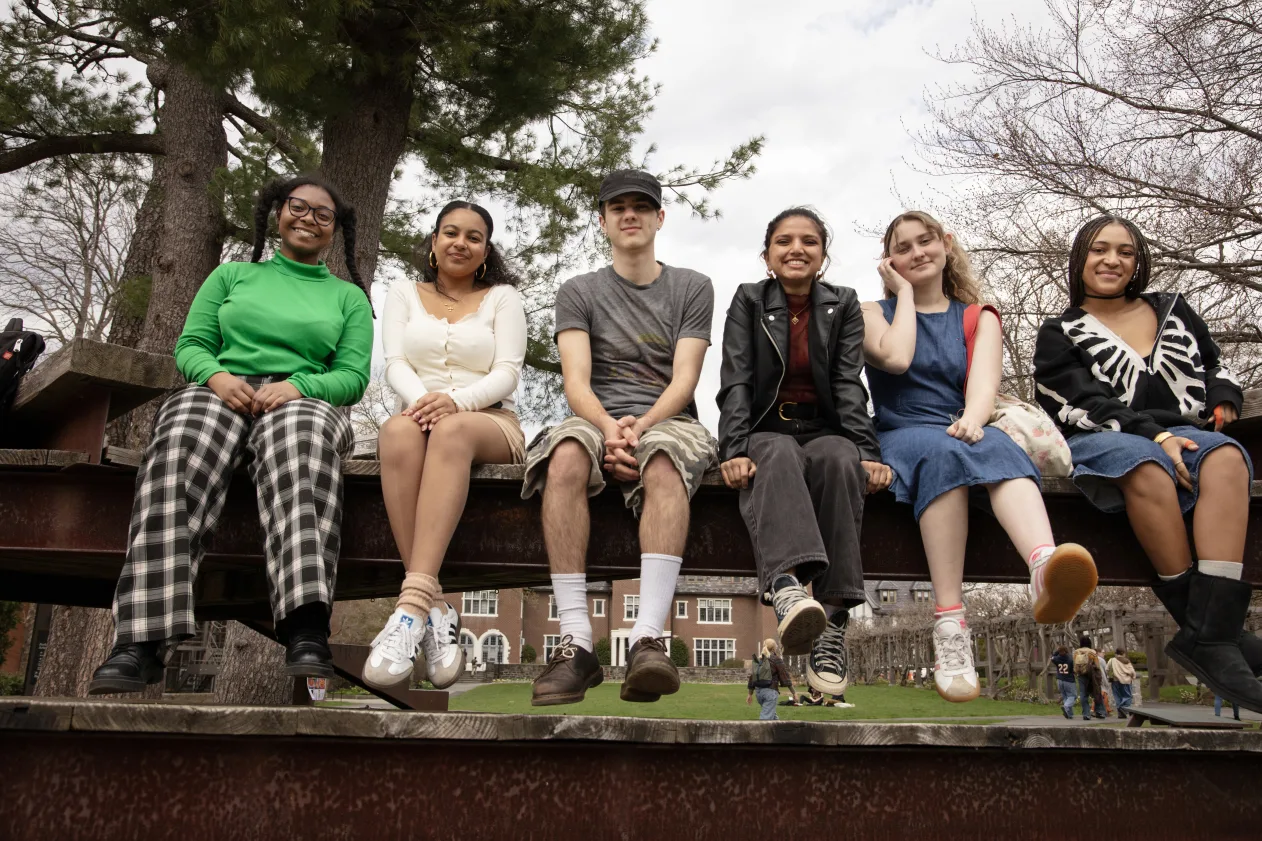 Six smiling people sit together on a wooden beam outside, with trees and a building in the background.