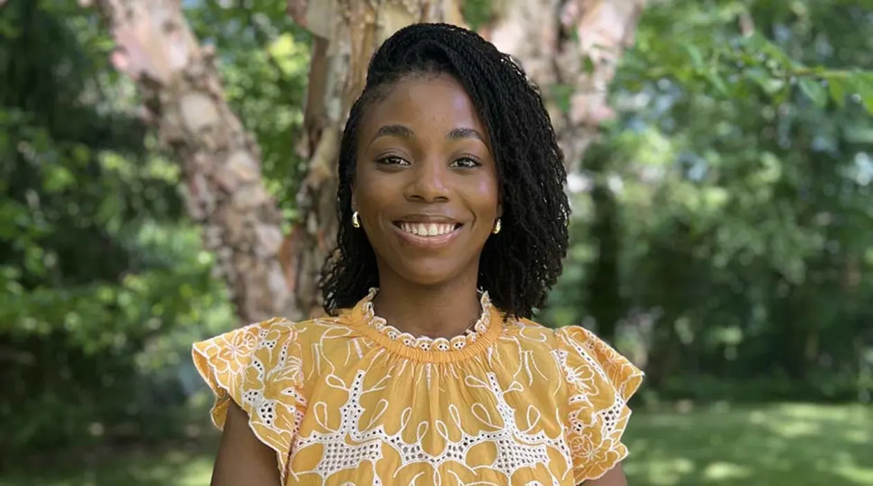 Person smiling outdoors, wearing a yellow patterned top. Trees and greenery are visible in the background.
