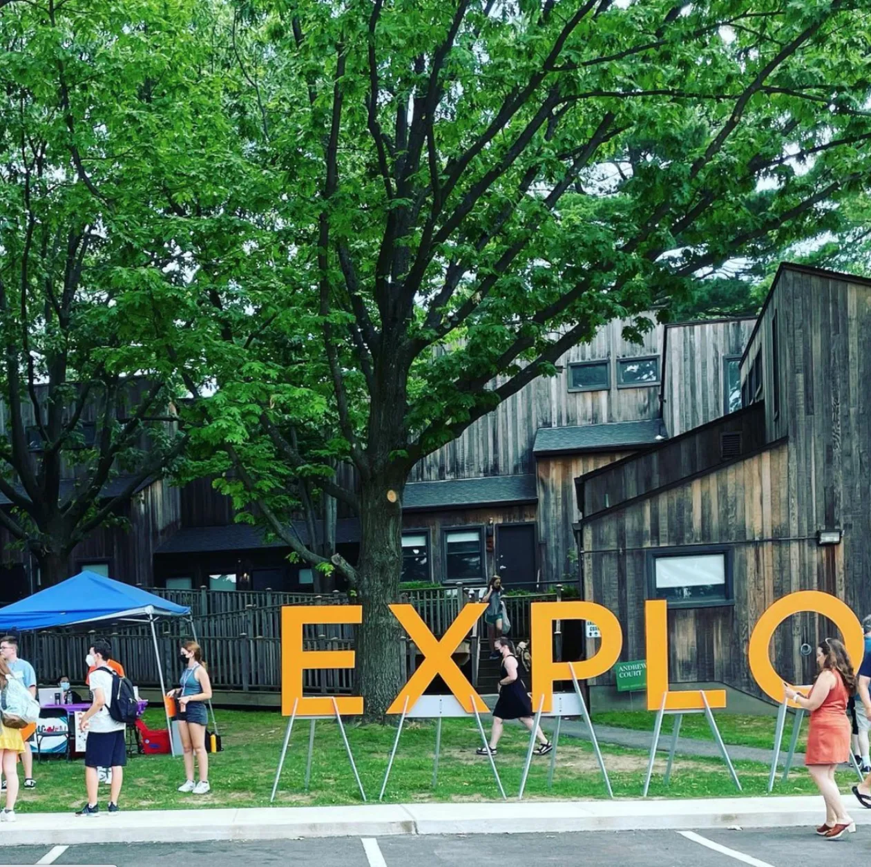 Large orange letters spelling "EXPLO" outdoors with people walking nearby, trees, and a rustic building in the background.